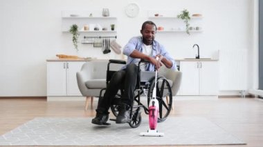 Mutfakta tekerlekli sandalyede otururken elinde kablosuz süpürge tutan engelli, yardımsever genç bir adam. Sıradan kıyafetli aktif kişi dairedeki halının tozunu ve pisliğini topluyor..