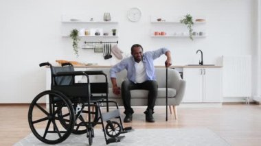 Afro-Amerikan tekerlekli sandalye kullanıcısının modern apartmanın açık-planlı mutfağında bastonla ayakta durmaya çalışmasının tam boy portresi..