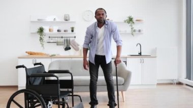 Engelli bir Afrikalı, tekerlekli sandalyeden kaldırırken baston kullanıyor. Açık planlı bir mutfağın ortasında..