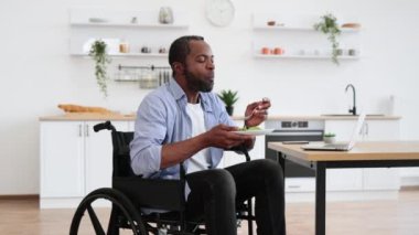 Tekerlekli sandalyedeki Afrikalı sakallı erkek mutfakta yemek sırasında bilgisayar kullanıyor. İşletme müdürü uzak bir ofiste kalırken tabakta vejetaryen salatasıyla enerjiyi artırıyor..