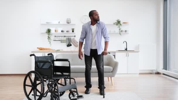 非洲残疾人在明亮的开放式厨房中间用手杖从轮椅上举起来 露出大拇指 — 图库视频影像