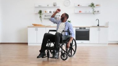 Modern bir apartmanın mutfağında dinlenirken gülümseyen ve dans eden Afro-Amerikan tekerlekli sandalye kullanıcısının tam boy portresi. Gündelik giysiler içinde genç bir erkek içeride zaman geçiriyor..