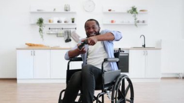 Engelli neşeli genç bir yetişkin modern apartman dairesinde joypad ile el ele tutuşuyor. Gülümseyen Afrikalı adam içeride kazananların jestleriyle bilgisayar oyunu oynuyor..