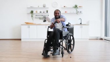 Engelli neşeli genç bir yetişkin modern apartman dairesinde joypad ile el ele tutuşuyor. Gülümseyen Afrikalı adam içeride kazananların jestleriyle bilgisayar oyunu oynuyor..