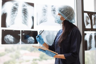 Muayene odasında tablet kullanırken siyah önlüklü ve tıbbi şapkalı bir kadın stilus kalemle vücut taramalarına bakıyor. Klinikteki tıbbi kayıtları analiz eden maskeli bir radyolog..