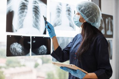 Yüz maskesi takan odaklanmış kadın doktor ve cam camına sabitlenmiş röntgen taramalarının yanında dijital tabletle poz veren üst fırça. Tıbbi eldivenli uzman Hindu cerrah yaraları inceliyor..