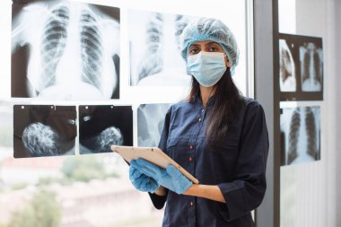 Muayene odasında tablet kullanırken siyah önlüklü ve tıbbi şapkalı bir kadın stilus kalemle vücut taramalarına bakıyor. Klinikteki tıbbi kayıtları analiz eden maskeli bir radyolog..