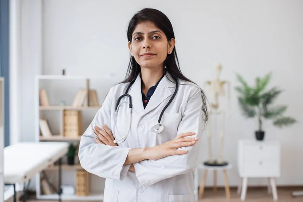 Qualified lady doctor in uniform standing confidently with crossed arms in modern clinic Interior. Reliable hindu specialist with stethoscope around neck representing professional medical skills.