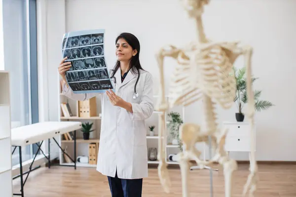 Confident indian doctor holding MRI scans of human brain in consulting room with skeleton model on blurred foreground. Professional female physician in medical uniform analyzing diagnostic report.