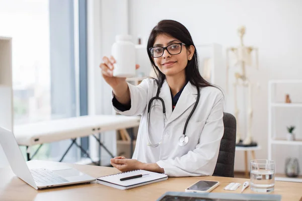 Component female doctor in indian nationality wearing lab coat and glasses sitting at desk with modern laptop and showing jar of pills at camera. Qualified advice about health treatment.