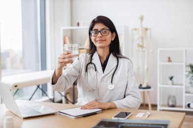 Beyaz laboratuvar önlüklü, dizüstü bilgisayarlı ve hastane ofisindeki belgelerle yazı masasında otururken bir bardak su gösteren pozitif Hintli kadın. Rekabetçi doktor susuz kalmamayı öneriyor..