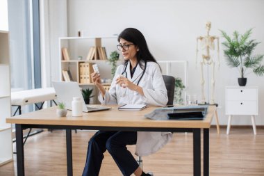 Dost canlısı bir kadın masada oturuyor ve elinde ilaç tutarken kamerayla hastayla sohbet ediyor. Hindu terapist ofisteki modern laptopu kullanarak tedavi öneriyor..