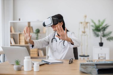 Laboratuvar önlüğü takan Hintli hemşire, dolabın bulanık arka planında modern cihazlarla masasında otururken VR gözlükleri takıyor. Kadın terapist, eğitim hastanesinde tıbbi eğitime hazırlanıyor..