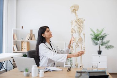 Pratisyen hekim insan iskeletinin modeline dokunuyor ve deftere kemiklerin yerini yazıyor. Hintli kadın doktor hastanede video dersi sırasında kişisel laptopuyla masada oturuyor..
