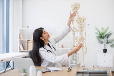 Steteskoplu kadın sıhhiyeci rahat iş yerinde iskelet anatomik modelini analiz ediyor. Video dersi sırasında kas iskeleti bozukluklarını açıklamak için dizüstü bilgisayar kullanan Hindu kadın..