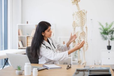 Kişisel dolabında iskelet modelinin yanında kişisel dizüstü bilgisayarı olan Hintli bir kadının yan görüntüsü. Esmer kadın doktor hastanede plastik kemiklere dokunuyor ve video çağrısı yapıyor..