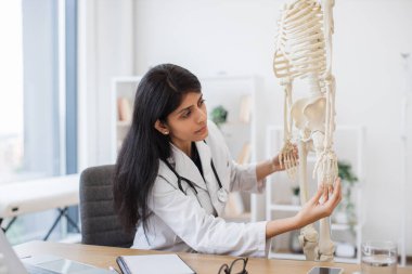 Becerikli bir kadın doktor, kişisel kabinede iş yerinde otururken plastik iskelet üzerinde el ele incelemesi yapıyor. Hintli başhekim insan vücudunun modelini kullanarak online konferans veriyor..