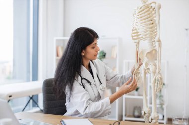 Becerikli bir kadın doktor, kişisel kabinede iş yerinde otururken plastik iskelet üzerinde el ele incelemesi yapıyor. Hintli başhekim insan vücudunun modelini kullanarak online konferans veriyor..