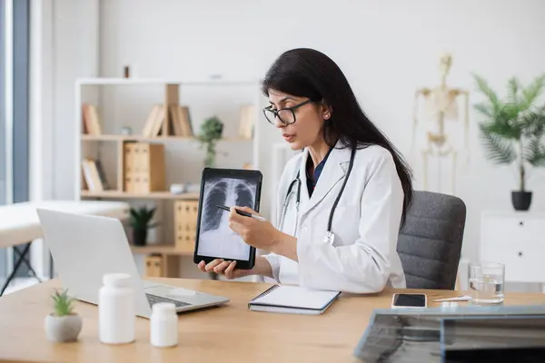 Side view of attractive nurse pointing on personal tablet with scan of x-rayed lungs. Kind female doctor having video call on modern laptop and talking about harmful formation in respiratory organs.