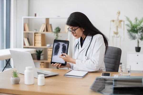 Side view of attractive nurse pointing on personal tablet with scan of x-rayed lungs. Kind female doctor having video call on modern laptop and talking about harmful formation in respiratory organs.