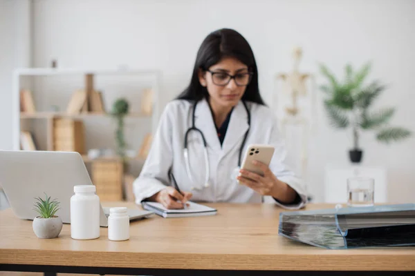 Attentive indian doctor sitting at table with smartphone and writing data of new patient into personal notebook. Indian dark haired woman in lab coat and glasses conducting video call for health care.