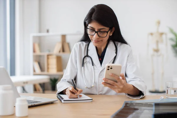 Serious medical practitioner wearing medical clothes using modern smartphone and taking notes during video call. Hindu female making consultation and prescribing treatment online.