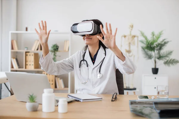 Female general practitioner holding hands up while wearing virtual reality glasses. Mature hindu woman in lab coat illustrating medical innovation technology in health center interior.