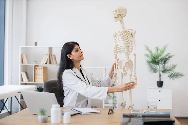 General practitioner touching model of human skeleton and writing location of bones in notebook. Indian female doctor sitting at table with personal laptop during video course in teaching hospital.