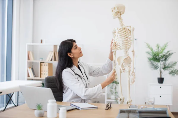 General practitioner touching model of human skeleton and writing location of bones in notebook. Indian female doctor sitting at table with personal laptop during video course in teaching hospital.