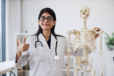 Özel bir klinikte, hastane önlüğü ve gözlük takan kadın doktorların insan vücudunun maketinin yanında durduğunu görmek. Çekici Hintli kadın modern işyerinde iskelet üzerinde pratik yapıyor, başparmağını kaldır.