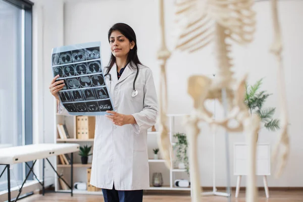 Confident indian doctor holding MRI scans of human brain in consulting room with skeleton model on blurred foreground. Professional female physician in medical uniform analyzing diagnostic report.