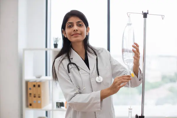 Adult female doctor in white lab coat standing near medical dropper and controlling liquid medication. Indian woman preparing infusion drip for intravenous treatment system.
