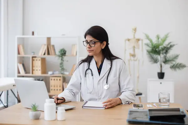Mature woman with stethoscope working in front of portable device at comfortable desk. Professional hindu doctor in white coat getting ready for online consultation in health center interior.