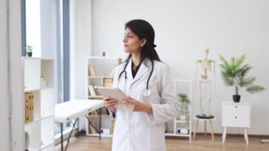 Doktorun ofisinde anatomik modelin arka planında poz verirken elinde tablet olan beyaz laboratuvar önlüklü Hindu kadın odaklanmış. İnsanlar, tıp ve teknoloji kavramı.