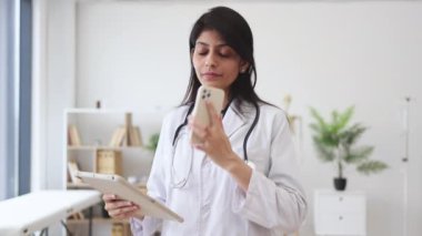 Tablet ekranına bakarken cep telefonuyla konuşan kadın doktorların ön görüntüsü. Yetenekli Hindu doktor, hastaneden modern cihazları iş ve danışmanlık için kullanıyor..