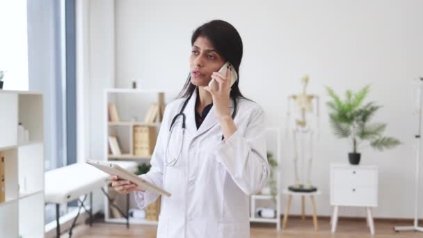 タブレットの画面を見ながら 女性医師が立って携帯電話で話す様子 病院から仕事や相談のために現代的なデバイスを使用して有能なヒンドゥー医師 — ストック動画
