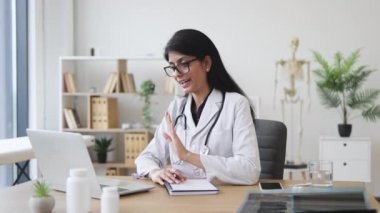Modern dizüstü bilgisayar kullanan uzman bir doktor ve online video konsültasyonu sırasında el kol hareketi yapıyor. Tıp üniformalı Hintli kadın, hasta bir hastaya masada otururken ev tedavisi planını anlatıyor..