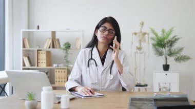Modern hastanedeki kişisel işyerinde hasta bir hastayla modern akıllı telefondan konuşan deneyimli Hintli doktor. Yetişkin bir kadın, hasta hakkında bilgi yazıyor..