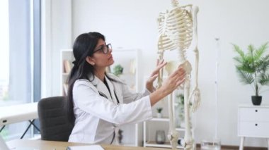Gözlüklü ve laboratuvar önlüklü pozitif hastane çalışanının portresi model iskeletin yanına oturmuş kameraya bakıyor. Olgun Hintli kadın muayene bozuklukları için koluna ve bacağına dokunuyor..