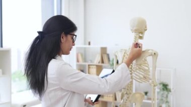 İskelet modelinin anatomisini açıklarken omurgasını gösteren tabletle Hindu tıp görevlisinin yan görüntüsü. Beyaz önlüklü kadın terapist hastane içinde insan vücudu yapısını inceliyor..