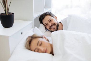 Gülümseyen sakallı koca, arka planda uyuyan karısı dinlenirken başının altında yastıkla rahatlıyor. Olgun bir ilişkisi olan neşeli, beyaz bir adam dolu bir hayattan zevk alıyor..