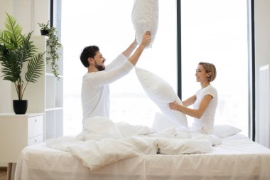 Genç tutkulu beyaz çift yastıklarla kavga ediyor, panoramik pencereli parlak yatak odasında uyandıktan sonra eğleniyorlar..