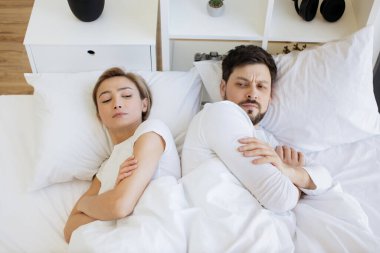 Beyaz yastığın üzerinde battaniyeyle uzanmış beyaz bir kadın ve kocası aydınlık yatak odasında uzanmış ona bakarken onu gücendirdi..