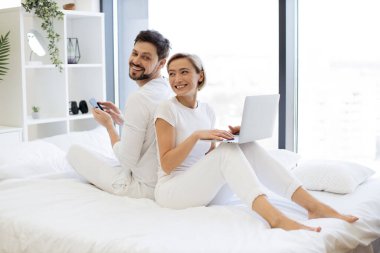 Genç bir kadın yatağında oturmuş modern dizüstü bilgisayarını kullanarak kısa mesajlar yollarken kocası da arkasında akıllı telefonuyla oturuyor. Evli çifti sevmek evde dinlenerek teknik yaşam dengesini sağlıyor..