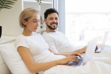 Yanındaki portatif bilgisayarda yazı yazmakla meşgul olan beyaz bir erkek dizüstü bilgisayarla yatakta sayfaları karıştırıyor. İlişkilerde olgun insanlar evde birlikte vakit geçirirler..