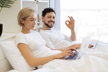 Beyaz erkek, arka planda taşınabilir bilgisayarda yazı yazmakla meşgulken yatakta dizüstü bilgisayardaki web sayfalarını karıştırıyor. İlişkileri olan insanlar evde birlikte vakit geçiriyorlar..