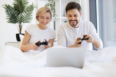 Evde battaniyenin altında yatarken beyaz bir çiftin oyun oynamalarının ön görüntüsü. Gülümseyen adam ve kadın, joystickleri kullanarak dizüstü bilgisayarda yarışırken eğleniyor..