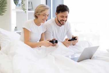 Beyaz bir çift evde battaniyenin altında yatarken video oyunu oynuyor. Gülümseyen adam ve kadın, joystickleri kullanarak dizüstü bilgisayarda yarışırken eğleniyor..