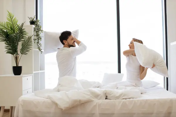 Young passionate Caucasian couple fighting with pillows, having fun after waking up in bright bedroom with panoramic windows.