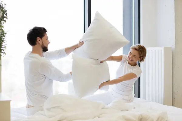 Side view of happy loving Caucasian husband and wife couple having pillow fight in bed, happily spending time on weekend.